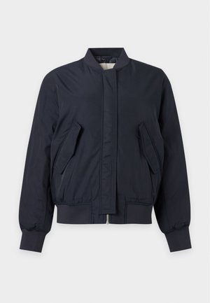 Γυναίκα που φοράει ένα σκούρο μπλε bomber μπουφάν, ανοιχτό γκρι τοπ, σκούρα, φαρδιά παντελόνια, μαύρες μπαλαρίνες, κρατώντας μια μαύρη τσάντα με τρύπες.
