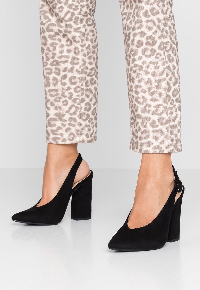 Escarpins slingback en suède noir avec bout pointu et motif découpé. Ils présentent un talon épais et un détail boucle près de la cheville.
