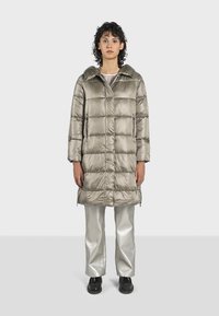 Cappotto imbottito grigio chiaro con colletto, caratterizzato da cuciture orizzontali e due tasche laterali. Indossato sopra pantaloni argentati lucidi e scarpe nere.