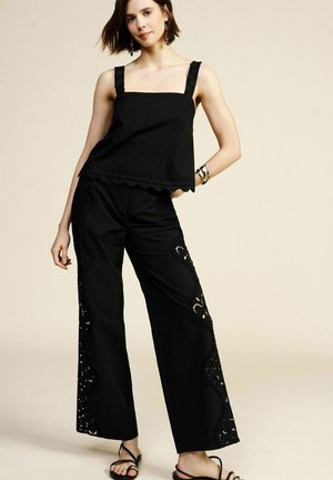 Giovane donna che indossa un top nero senza maniche e pantaloni neri a gamba larga con ritagli in pizzo, in piedi davanti a uno sfondo neutro.