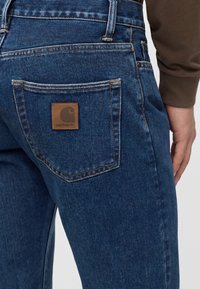 Detailní záběr kapsy modrých džínů s hnědou nášivkou loga Carhartt, kterou nosí osoba s hnědým tričkem s dlouhým rukávem.