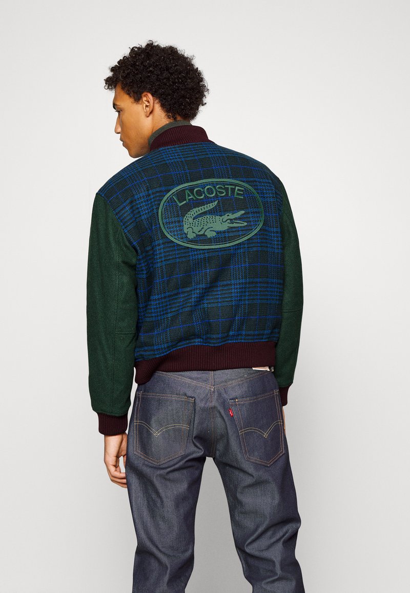 Veste varsity avec un corps vert foncé, motif à carreaux bleus, col et poignets côtelés, et logo Lacoste brodé au dos.