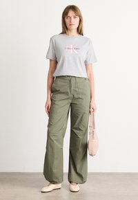 Camiseta de algodón gris con el logo de 'Calvin Klein', combinada con pantalones holgados de color verde oliva. El modelo sostiene una bolsa de color beige claro. Fondo blanco.