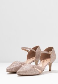 Chaussures à talons hauts en rose doré avec un bout pointu, une bride de cheville et un tissu texturé. Petit talon et design de bride croisée. Doublure intérieure lisse.