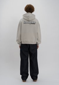 Felpa oversized beige con cappuccio regolabile, caratterizzata da un logo distintivo sul retro. Abbinata a jeans a gamba larga blu scuro.