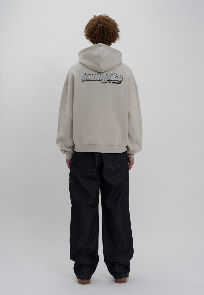 Felpa oversized beige con cappuccio regolabile, caratterizzata da un logo distintivo sul retro. Abbinata a jeans a gamba larga blu scuro.