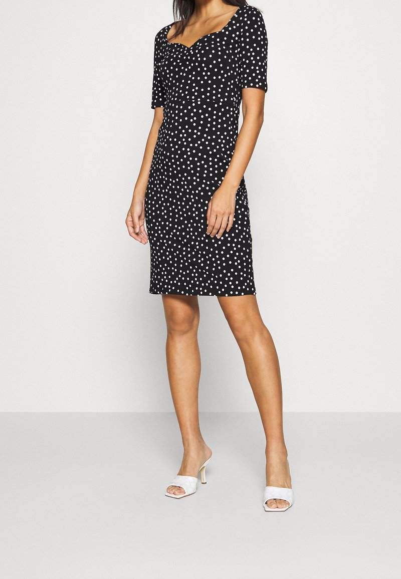 Femme portant une robe noire mi-longue à pois blancs et manches courtes, associée à des sandales blanches à talons hauts et sans brides.