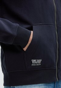 Sudadera con cremallera en color navy, con puños y dobladillo acanalados, bolsillos frontales y una etiqueta de Camp David con texto en la esquina inferior izquierda.