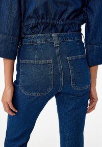 Mörkblå denimjeans med hög midja, med två bakfickor och synliga sömnadsdetaljer. Texturen är något strukturerad och hållbar.