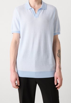 Mand iført en lyseblå kortærmet strikket polo med sorte bukser, der viser en tatoveret højre underarm.