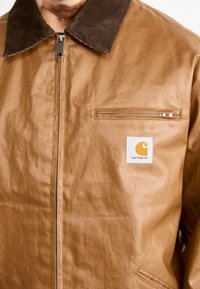 Giacca marrone con texture, colletto in corduroy scuro, zip frontale e una piccola etichetta bianca del marchio Carhartt sul taschino del petto.