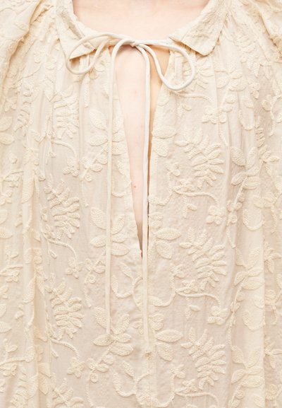 Κρουασάν φόρεμα με κέντημα σε κρεμ απόχρωση, που διαθέτει βαθύ V-λαιμόκοψη και κλείσιμο με δέσιμο. Το ύφασμα είναι υφασμένο με λουλουδένια σχέδια και φουσκωτά μανίκια.