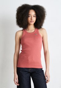 Röd ribbad linne med rund halsringning och en liten grön Lacoste-logga på bröstet, kombinerat med mörka denimbyxor. Jämn struktur genomgående.