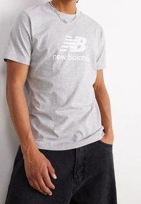 Persona vistiendo una camiseta gris de New Balance, un collar de cadena plateada y pantalones negros, con una mano en el bolsillo, frente a un fondo blanco.