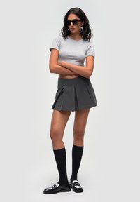 T-shirt grigio corto, gonna plissettata grigio scuro, calze nere fino al ginocchio, mocassini neri lucidi con accenti bianchi, modella con capelli ricci e occhiali da sole.