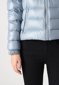 Fôret lys blå jakke med glidelåslomme, blank finish og trykknappmansjetter, sammen med svarte jeans for et avslappet antrekk.