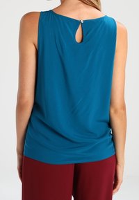 Haut sans manches bleu sarcelle avec un détail dos en forme de goutte orné d'un petit bouton. Tissu lisse avec une coupe décontractée. Associé à un pantalon bordeaux.