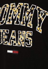 Svart bomulls t-shirt med vit text "TOMMY" och "JEANS", accentuerad med gula blommönster. Litet randigt logodetalj nedanför.