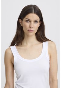 Sieviete ar gariem brūniem matiem, tērpusies baltā bezroku topā, skatās tieši kamerā ar neitrālu izteiksmi uz vienkārša fona.