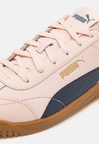 Svetle růžová teniska Puma s námořnickým bočním pruhem, béžovou podrážkou, odpovídajícími růžovými tkaničkami a zlatým logem Puma na boku.