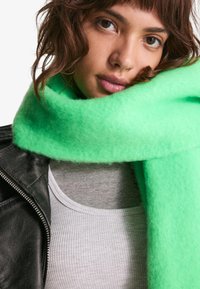 Zelena volnena šala z mehko, puhasto teksturo ovita okoli vratu, v kombinaciji s sivo rebrasto majico in črnim usnjenim jopičem.