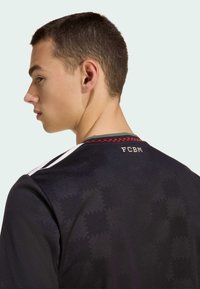 Young man with short hair wearing a black shirt with subtle checkered pattern and "FCBM" printed at the back collar.