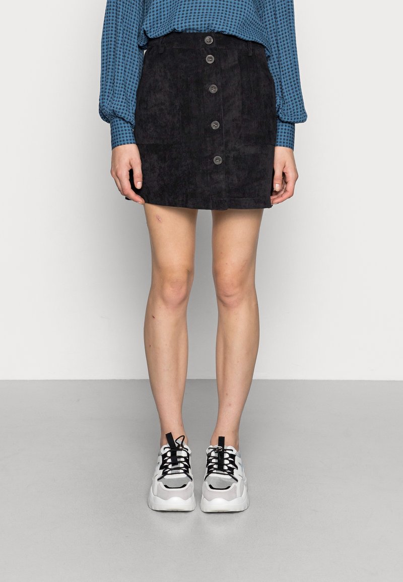 Personne portant une mini-jupe noire à boutons sur le devant, un haut bleu à manches longues avec motif et des baskets blanches, debout sur un sol gris contre un mur blanc.