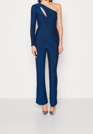 Mono con un hombro en tejido azul oscuro brillante, que presenta un detalle recortado y pantalones acampanados, combinado con tacones morados transparentes.