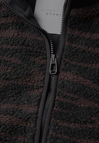 Gros plan d'une veste polaire texturée noire et marron avec une fermeture éclair noire partiellement fermée et une étiquette blanche à l'intérieur indiquant "STREET STUDI".