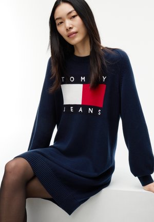 Ung kvinde med langt mørkt hår iført en navyblå Tommy Jeans sweaterkjole, der sidder med det ene ben krydset.