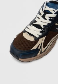 Αθλητικά παπούτσια με καφέ διχτυωτό άνω μέρος και μαύρες και κρεμ δερμάτινες λεπτομέρειες. Κρεμ κορδόνια, μπλε λεπτομέ ρεια κοντά στο δάχτυλο του ποδιού και μια υφασμάτινη καφέ σόλα.