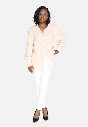 Lyse ferskenfarvet skjorte med knappelukning og tekstureret stof samt krave, parret med hvide skinny jeans og sorte højhælede sko. Simpelt design og neutrale farver.