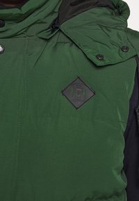 Vihreä toppatakki, jossa on korkea kaulus, mustat yksityiskohdat, mattamustat napit ja timantinmuotoinen logopatch rinnassa.