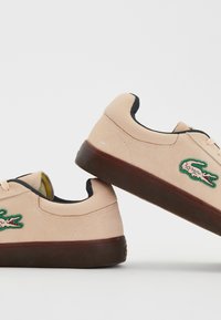 Paire de baskets en daim beige avec semelles marron foncé et logo crocodile vert sur le côté, sur fond blanc.