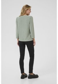 Haut vert clair avec texture, à manches trois-quarts et encolure arrondie, associé à un pantalon noir slim et des chaussures noires.