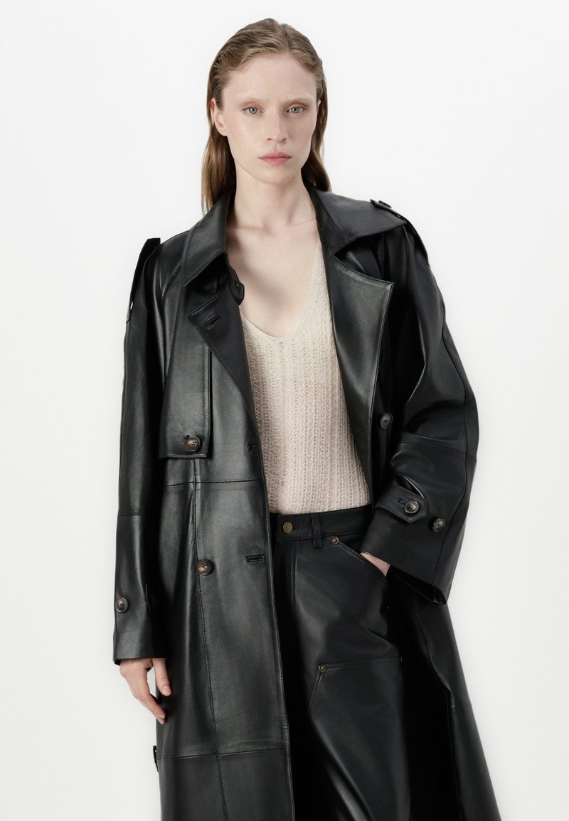 Schwarzer Leder-Trenchcoat mit doppelter Knopfleiste, breiten Revers und Seitentaschen, kombiniert mit einem hellen Strickoberteil und dunklen Hosen.