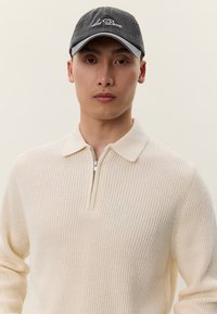 Junger Mann trägt einen cremefarbenen, gerippten Half-Zip-Pullover und eine dunkelgraue Baseballkappe mit weißem, gesticktem Text.