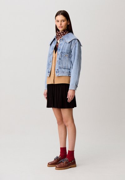 Veste en jean à grand col, de couleur bleu clair ; portée sur un cardigan beige, une jupe plissée noire, des chaussettes rouges et des chaussures marron avec des accents.