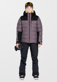 Lille puffy jakke i lilla og sort med lynlås, featuring hætte og kontrasterende ærmer; sorte skibukser; holder sorte handsker; skibriller på hovedet.