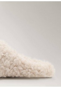 Pantofole morbide di un bianco sporco con una superficie soffice e testurizzata e un bordo arrotondato, caratterizzate da un design minimalista e senza hardware visibile.