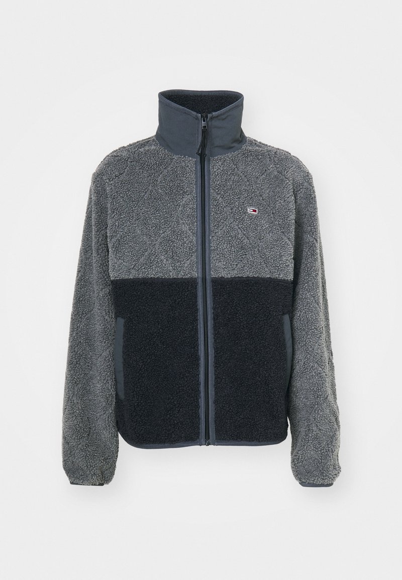 Grå og sort fleecejakke med høj krave, lynlås foran, struktureret krop og logodetalje. Har blødt stof og kontrasterende farveblokke.