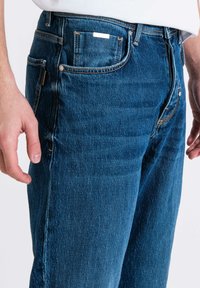 Jeans in denim blu con taglio dritto, dotati di cinque tasche, cuciture decorative visibili e una piccola etichetta del marchio sulla vita.
