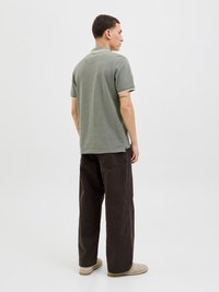 Krátký rukáv, zelený polo s kontrastním světlým límcem a manžetami, spárovaný s volně padnoucími tmavě hnědými kalhotami a béžovými botami.