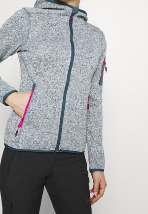 Osoba ubrana w szarą bluzę z kapturem zapinaną na zamek z różowymi akcentami na zamku oraz czarne spodnie, z ręką umieszczoną w kieszeni bluzy.