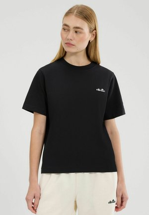 Kobieta o długich blond włosach, nosząca czarny t-shirt marki ellesse i kremowe szorty ellesse, patrząca w lewo na jednolitym tle.