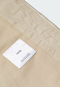 Tessuto beige con cuciture visibili e un'etichetta bianca con la scritta "MNG" e informazioni sulle cure, posizionata all'interno di un indumento.