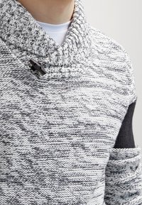 Stickad tröja med polokrage, ett strukturerat mönster i grått och vitt, samt en svart accent på vänster axel. Inkluderar en knapppå detalj.