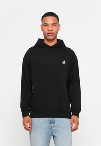 Schwarzes Kapuzensweatshirt mit Fronttasche, gerippten Bündchen und Saum. Mit einem kleinen vielfarbigen Dreieckslogo auf der linken Brust.