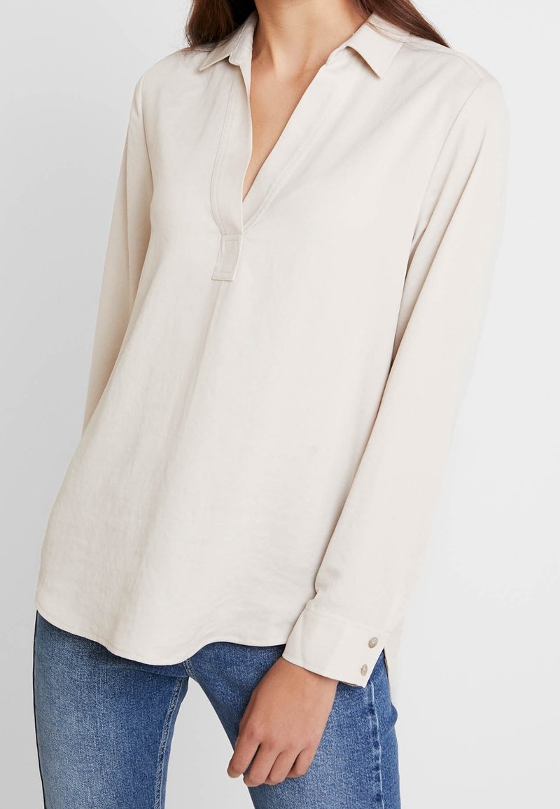 Blouse beige clair à manches longues avec col en V et petite patte de boutonnage, confectionnée dans un tissu doux, avec des manches à revers ornées de boutons.