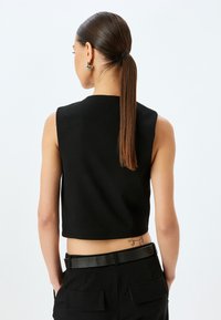 Schwarzes, kurzes, ärmelloses Oberteil mit einem klaren Design, das einen hohen Kragen und eine glatte Textur aufweist, kombiniert mit hochtaillierten Hosen und einem Gürtel.